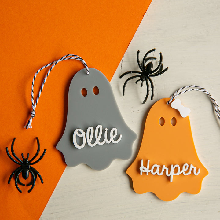 Two ghost ornaments, one grey with "Ollie" and one orange with "Harper", feature string loops and personalised Acrylic Halloween Tags. Two black spiders are placed nearby on white and orange backgrounds.
