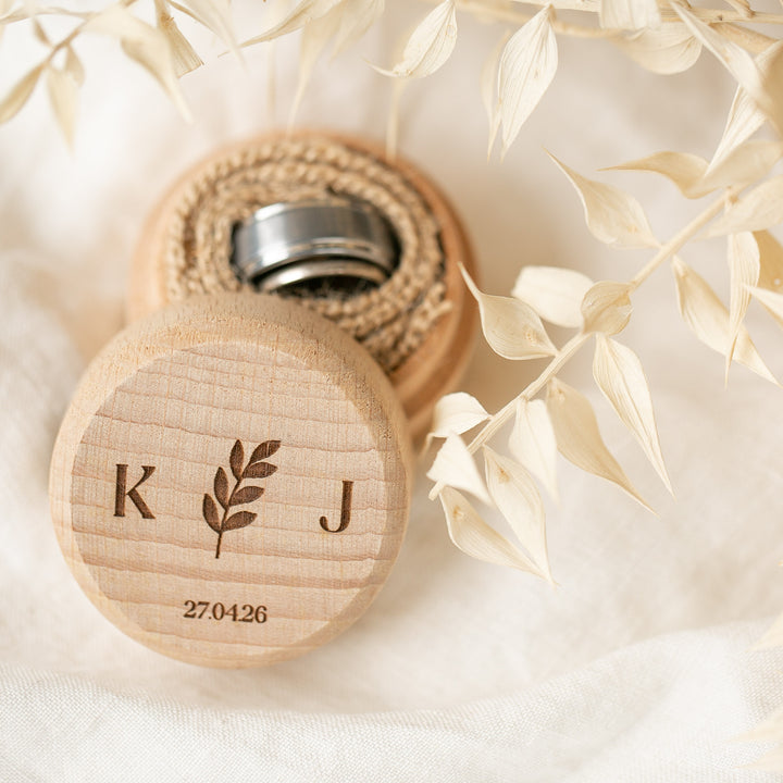 A Personalised Ring Box with a lid engraved with “K,” a leaf, “J,” and the date “27.04.26” sits open, displaying two silver rings inside amid dried pale leaves on soft white fabric.