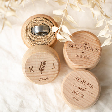 Three Personalised Ring Boxes rest on cream fabric, two engraved with names, initials, and dates. One wooden box is open to reveal a ring inside on burlap, with dried white foliage.