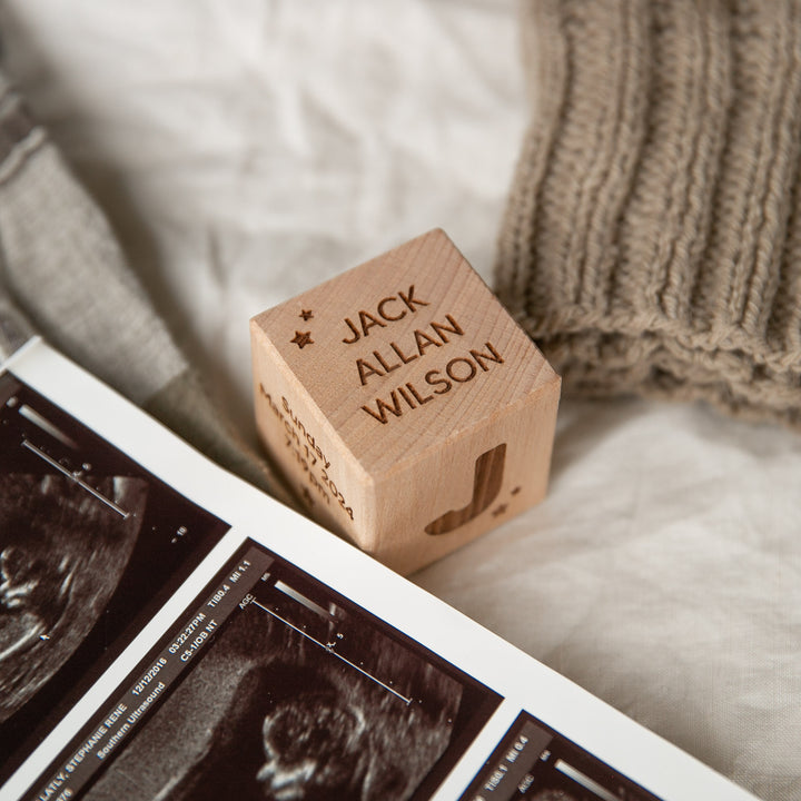 A Personalised Wooden Block engraved with a name sits on a soft surface beside knitted fabric and ultrasound images, making it a charming baby keepsake—perfect for announcements or celebrations.