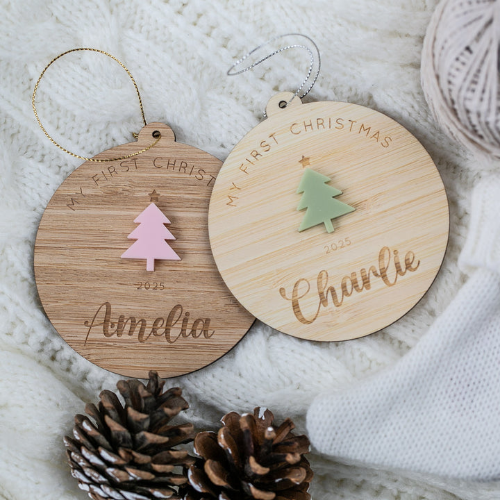 Two wooden Christmas ornaments with tree designs and engraved names on a soft fabric background.