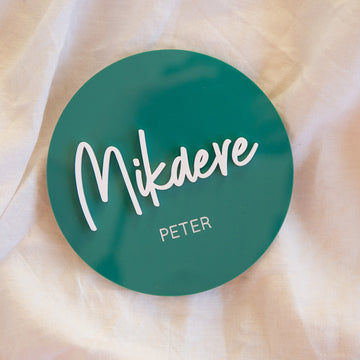 A round green acrylic plaque featuring "Mikaere" handwritten in white and "Peter" printed below, set on cream fabric—ideal as a Personalised Acrylic Name Announcement or custom nursery decor.