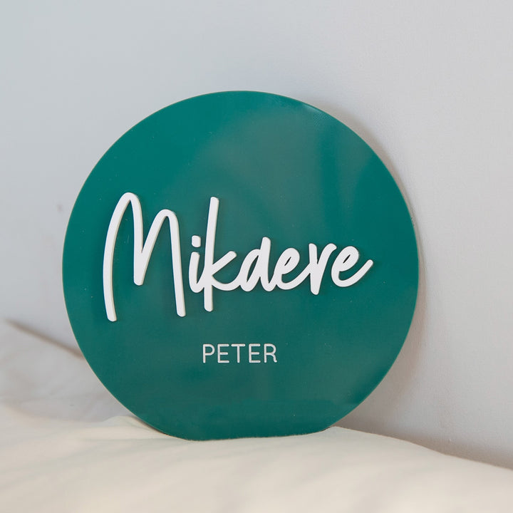 A round, teal Personalised Acrylic Name Announcement rests against a white surface. "Mikaere" appears in large white cursive, with "PETER" in smaller uppercase below—ideal for custom nursery decor.
