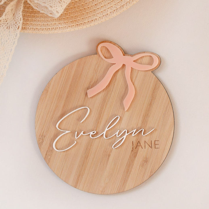 The Personalised Bow Plaque is a round wooden name sign with a pink bow, featuring the name Evelyn in white script and Jane in smaller capitals—ideal for girls’ room decor or as a unique baby shower gift.
