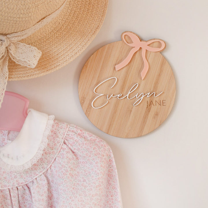 The Personalised Bow Plaque—a round wooden name sign with a pink bow and white script—adds sweet charm to a girl&