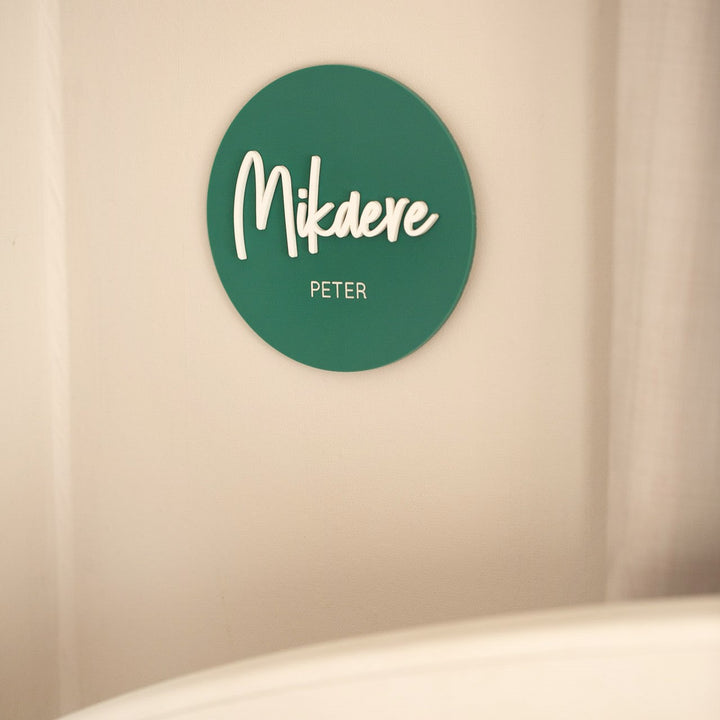 A green circular Personalised Acrylic Name Announcement is displayed on a light wall, featuring "Mikdere" in large white script and "Peter" below. Part of a white object is visible at the bottom, making it perfect custom nursery decor.