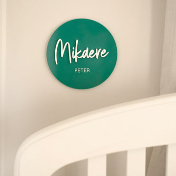 A green circular "Personalised Acrylic Name Announcement" plaque on the wall displays Mikdere in large script and Peter in smaller print, creating custom nursery decor above a white crib rail.