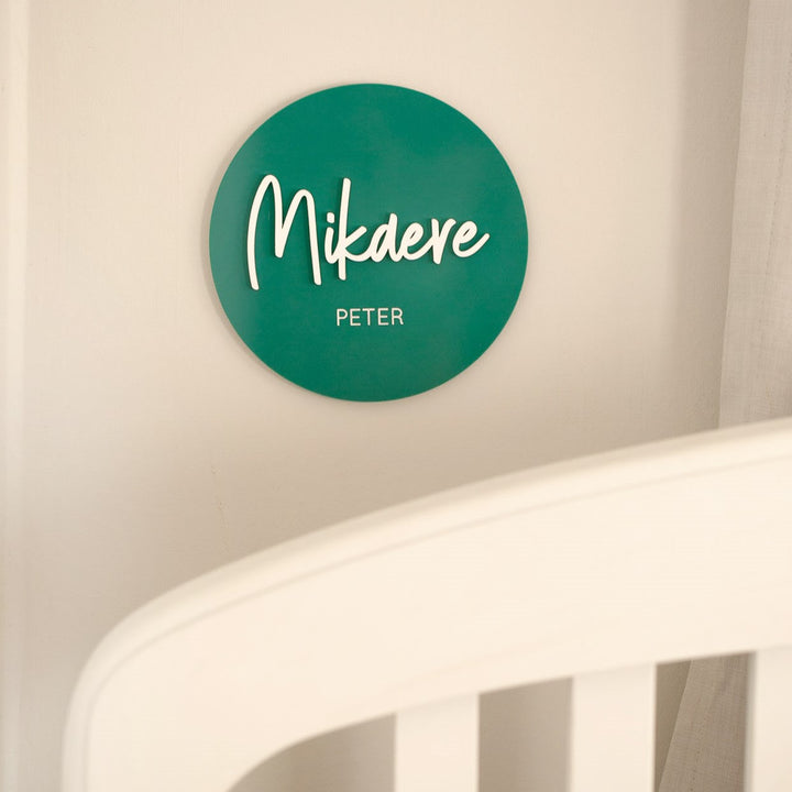 A green circular "Personalised Acrylic Name Announcement" plaque on the wall displays Mikdere in large script and Peter in smaller print, creating custom nursery decor above a white crib rail.