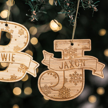 Two Jolly Alphabet Christmas Baubles, “B” and a personalised “J” for Jaxon, hang on a tree. Each features snowflake engravings and a ribbon banner, with blurred festive lights twinkling in the background.