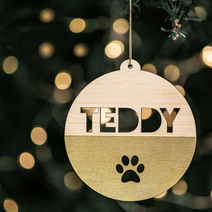 A Personalised Pet Christmas Bauble hangs on a tree, made of wood with gold glitter. It features TEDDY and a paw print cutout, glowing warmly against softly blurred festive lights in the background.