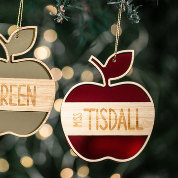 Two apple-shaped “Apple for the Teacher Christmas Bauble” ornaments—one gold “GREEN” and one red and white “MISS TISDALL”—hang from a tree, surrounded by blurred lights and pine needles in the background.