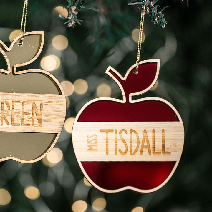 Two apple-shaped “Apple for the Teacher Christmas Bauble” ornaments—one gold “GREEN” and one red and white “MISS TISDALL”—hang from a tree, surrounded by blurred lights and pine needles in the background.