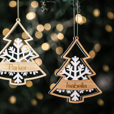 Two Frosted Forest Christmas Baubles with white snowflake designs hang on a tree, each personalised, Parker on the left and Isabella on the right. Blurred lights in the background.