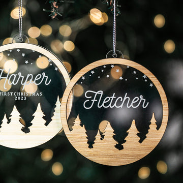 Two Snow Globe Christmas Baubles with forest and star designs hang on a tree—one engraved with “Harper First Christmas 2023,” the other with “Fletcher.” Soft twinkle lights blur in the background.