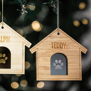 Two Furry Friends Christmas Bauble ornaments, shaped like wooden dog houses with paw print cutouts, hang from a tree. Each is personalised—RALPH on the left, TEDDY on the right—with blurred festive lights in the background.