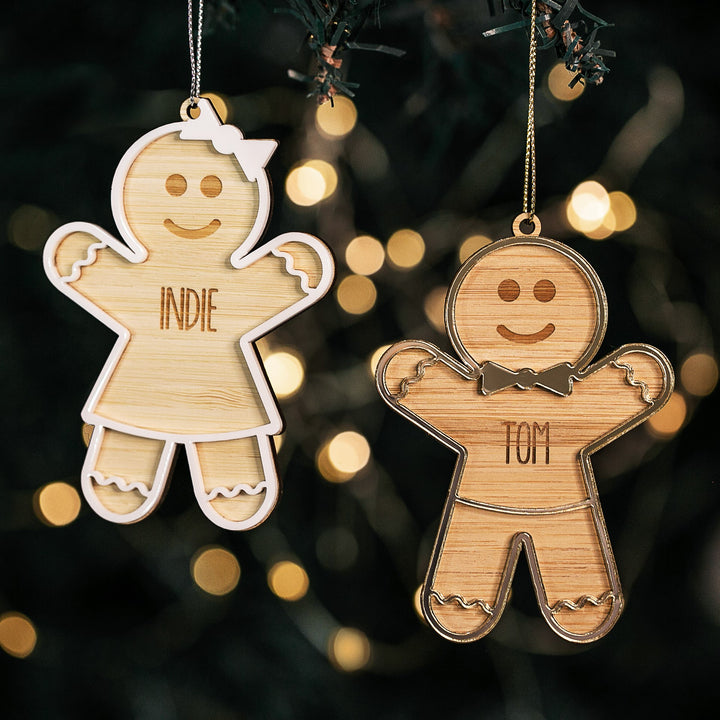 Two Gingerbread Christmas Bauble ornaments—one personalized "INDIE" with a bow and dress, the other "TOM" with a bow tie—hang from a Christmas tree, softly glowing among blurred festive lights.