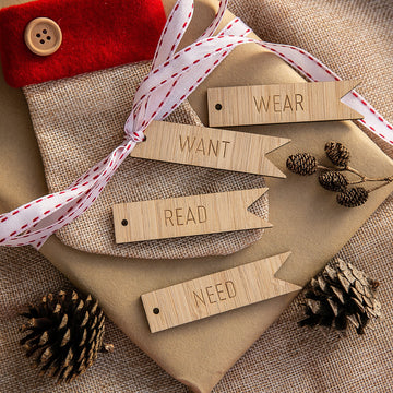 The unpersonalised Flag - Want, Need, Wear, Read Tags are arranged on wrapped gifts with white and red ribbon, pinecones, and a fabric pouch with a button atop a burlap background.