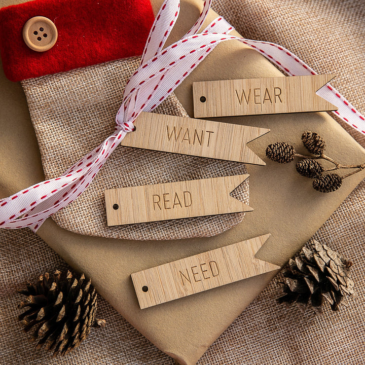 The unpersonalised Flag - Want, Need, Wear, Read Tags are arranged on wrapped gifts with white and red ribbon, pinecones, and a fabric pouch with a button atop a burlap background.