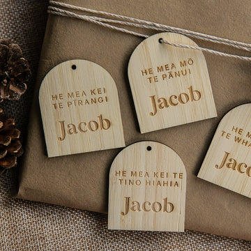 Four Personalised Te Reo - Want, Need, Wear, Read Tags featuring the name Jacob are arranged on a wrapped gift with twine and pinecones on burlap.