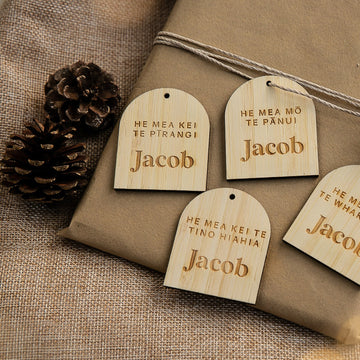 Four wooden "Personalised Te Reo - Want, Need, Wear, Read Tags" engraved with the name Jacob and Māori phrases are displayed on a brown wrapped gift with twine and pinecones.