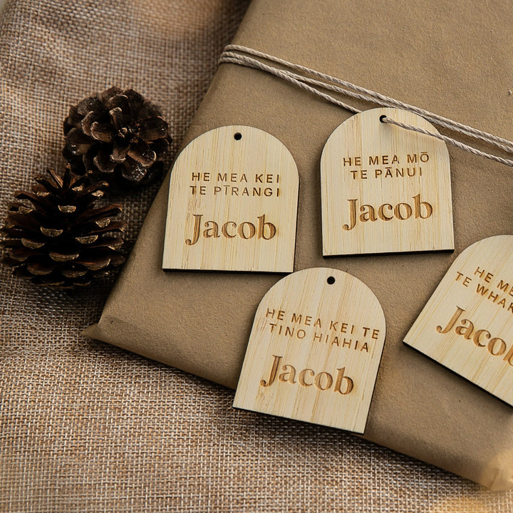 Four wooden "Personalised Te Reo - Want, Need, Wear, Read Tags" engraved with the name Jacob and Māori phrases are displayed on a brown wrapped gift with twine and pinecones.