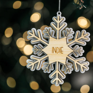 The Snow Crystal Christmas Bauble, a custom wooden snowflake ornament engraved with "INDIE," hangs on a Christmas tree amid blurred golden lights in the background.
