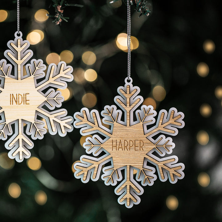 Two Snow Crystal Christmas Baubles hang on a Christmas tree, each engraved with a name—one says INDIE and the other HARPER. These personalized ornaments stand out against softly blurred holiday lights.