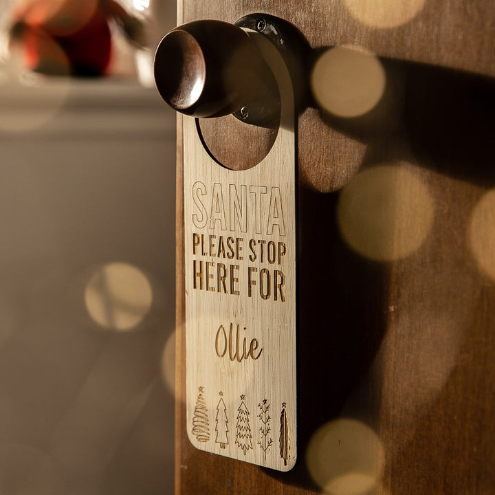 The Santa Stop Here Door Hanger reads “SANTA PLEASE STOP HERE FOR Ollie” with small Christmas trees at the bottom. Warm festive lighting creates a cozy backdrop, making this a charming personalized Christmas tradition.