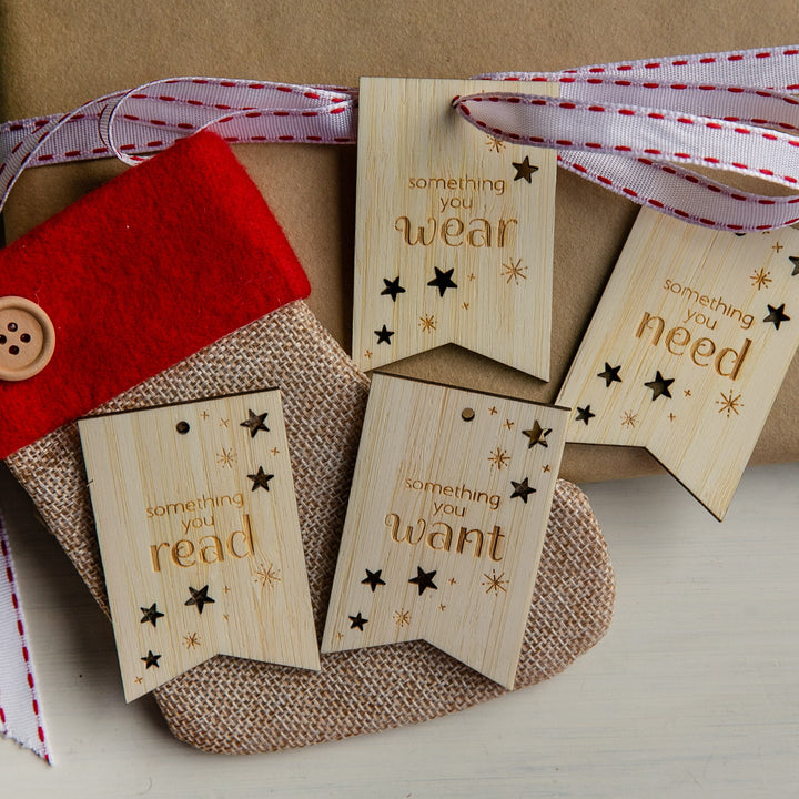 A burlap and red felt Christmas stocking with a button sits beside a Twinkle Twinkle Want, Need, Wear, Read Tags set—three wooden Christmas gift tags—on a ribbon-wrapped present.