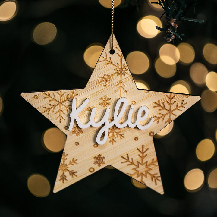 The Northern Star Christmas Bauble, a wooden ornament engraved with snowflake designs and the name Kylie in white letters, hangs on a tree amid softly glowing, blurred background lights.