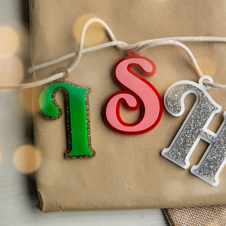 A close-up of a wrapped gift topped with Personalised Deluxe Letter Gift Tags in S, H, and I shapes (red, white, and green), strung together on a white cord.