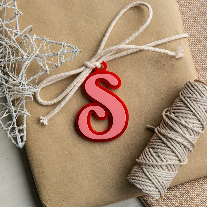 A wrapped gift with a white string bow, decorated with a red Personalised Deluxe Letter Gift Tag and a silver wire star, sits beside a roll of twine on a textured surface.