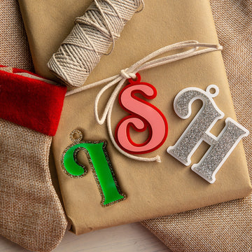 A gift wrapped with twine features three Personalised Deluxe Letter Gift Tags, "S" in red, "H" in silver glitter, and "i" in green with gold trim, next to a burlap stocking with a red cuff.
