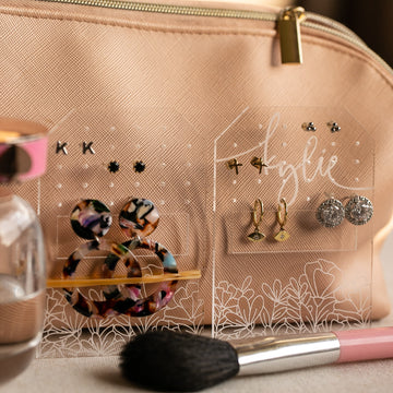 The Personalised Travel Earring Holder displays colorful hoops and studs in front of a beige makeup bag, with a makeup brush and perfume bottle nearby—an ideal organizer for your jewellery on the go.