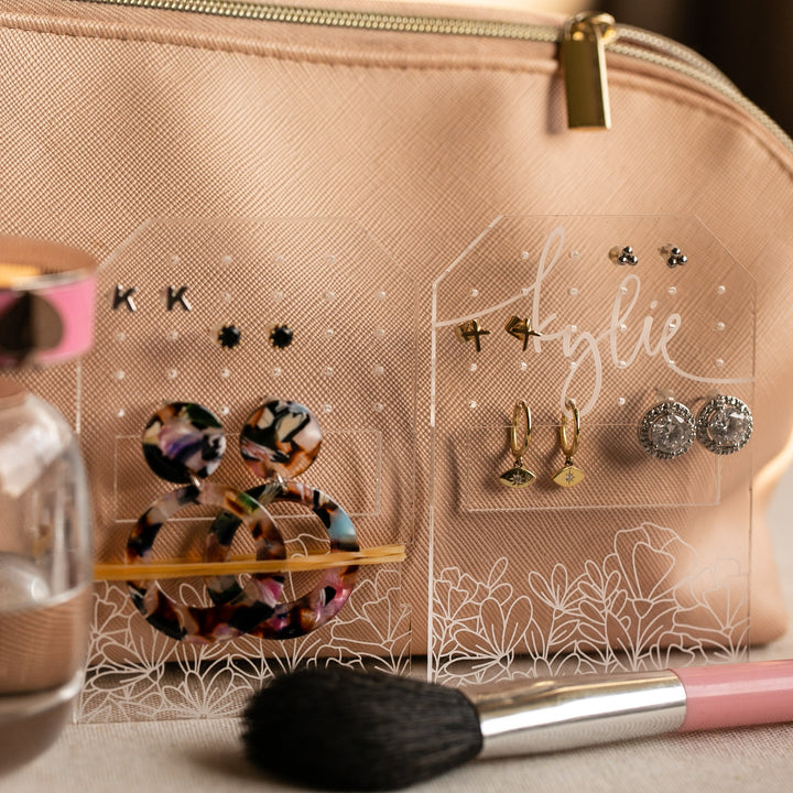 The Personalised Travel Earring Holder displays colorful hoops and studs in front of a beige makeup bag, with a makeup brush and perfume bottle nearby—an ideal organizer for your jewellery on the go.