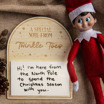 The Personalised Note from Elf on the Shelf features a festive elf doll beside a wooden sign, with a custom message board that reads, “Hi! I’m here from the North Pole to spend Christmas with you.” Pinecones and burlap decorate the background.