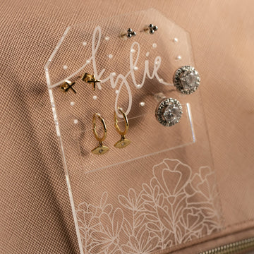 The Personalised Travel Earring Holder, made of clear acrylic and labeled with "kylie," displays five pairs of gold and silver earrings—studs, hoops, and large rounds—with a floral design etched at the base. Ideal for organizing jewellery on the go.