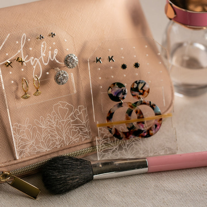 A makeup brush sits on a beige makeup bag next to a Personalised Travel Earring Holder showcasing earrings, with a small glass bottle topped by a pink lid.