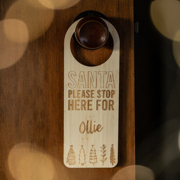 The Santa Stop Here Door Hanger, personalised with "Santa please stop here for Ollie" and engraved Christmas trees at the bottom, adds a charming touch to your festive tradition when hung on a brown door.