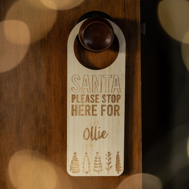 The Santa Stop Here Door Hanger, personalised with "Santa please stop here for Ollie" and engraved Christmas trees at the bottom, adds a charming touch to your festive tradition when hung on a brown door.