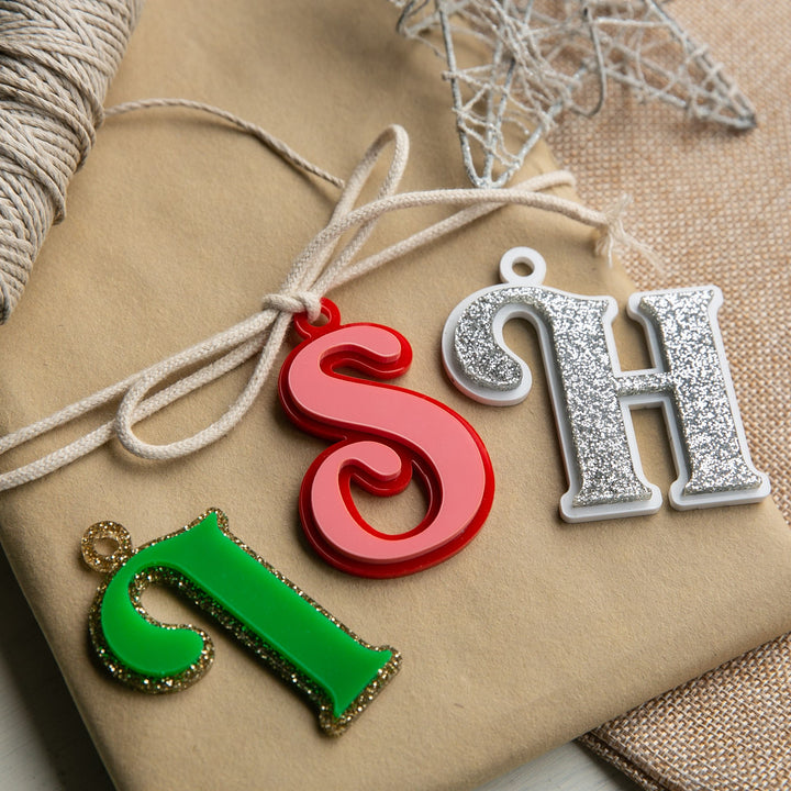 Three decorative letter ornaments, an ornate green J, shiny red S, and glittery silver H, rest on tan paper with twine and a silver star. Personalised Deluxe Letter Gift Tags.