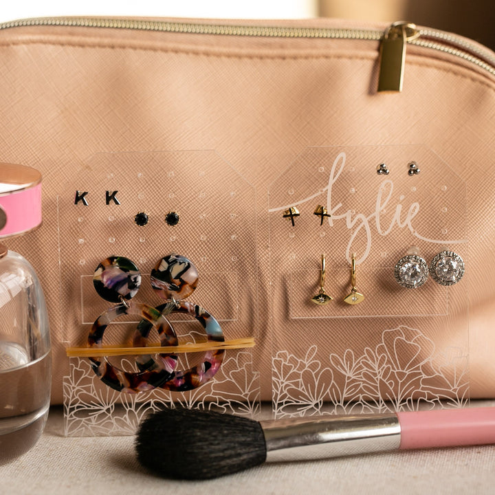 A Personalised Travel Earring Holder is displayed in a beige makeup bag with a gold zipper, surrounded by various earrings, a makeup brush, and a pink perfume bottle on a wooden surface.