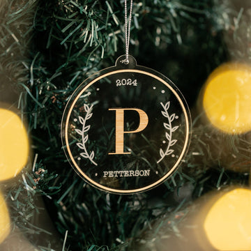 A round Christmas bauble with a gold monogram in the center, surrounded by green foliage and the year 2024 engraved at the top. The bauble is hanging from a Christmas tree with blurred lights in the background.