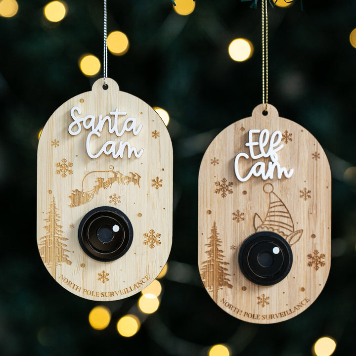 Two Santa Surveillance Christmas Bauble ornaments shaped like cameras hang on a tree. One reads "Santa Cam" with a reindeer and sleigh; the other says "Elf Cam" with an elf hat. Both feature festive snowflakes and faux camera lenses.