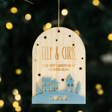 The "Our First Home Christmas Globe" hangs on a tree, engraved with “ELLY & CHRIS, Our First Christmas at 25 Smith Road, 2024,” and features house and tree cutouts plus star-shaped holes, with blurred festive lights in the background.