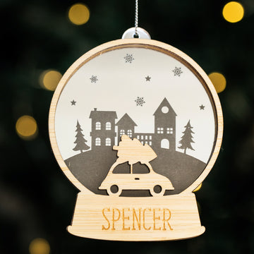 The Personalised Christmas Snowglobe is a wooden ornament shaped like a globe, featuring a car with a tree on top and "SPENCER" engraved at the base—a unique festive decoration for your Christmas tree.