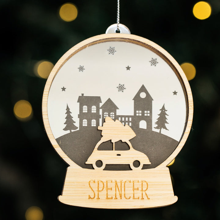 The Personalised Christmas Snowglobe is a wooden ornament shaped like a globe, featuring a car with a tree on top and "SPENCER" engraved at the base—a unique festive decoration for your Christmas tree.