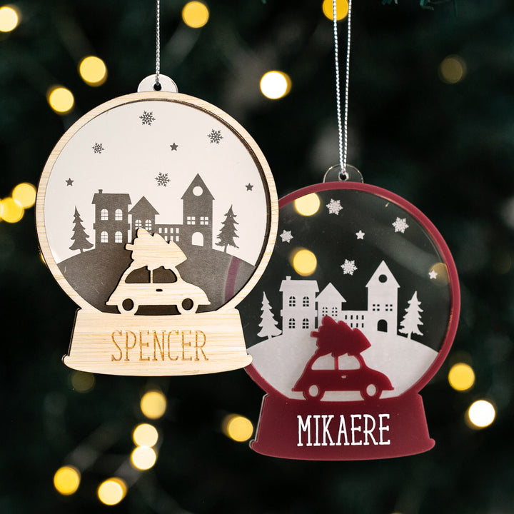 Two personalised Christmas snow globes with a car and a Christmas tree design, hanging from holiday lights.