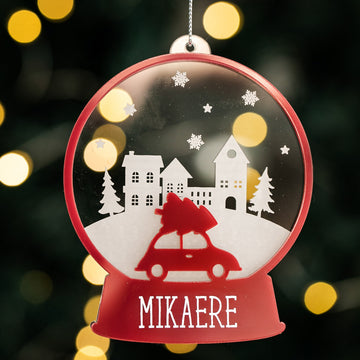 The Personalised Christmas Snowglobe is a red bauble shaped like a snow globe, featuring a car with a tree, snowy houses, pine trees, snowflakes, and the name Mikaere at the base—a charming festive tree decoration.