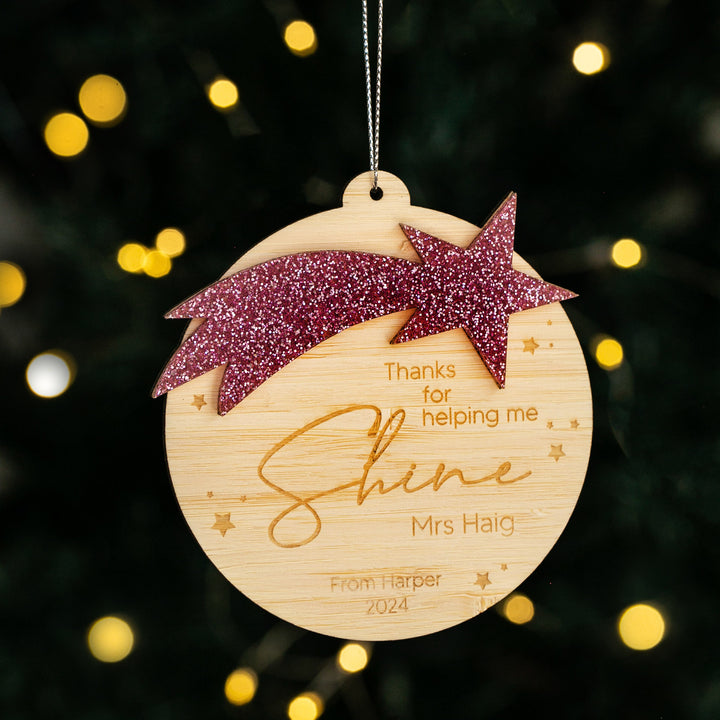 The Shining Star Teacher Christmas Bauble is a wooden ornament with a glittery pink shooting star and engraved text: Thanks for helping me shine Mrs Haig. From Harper 2024. Blurred Christmas lights glow in the background.